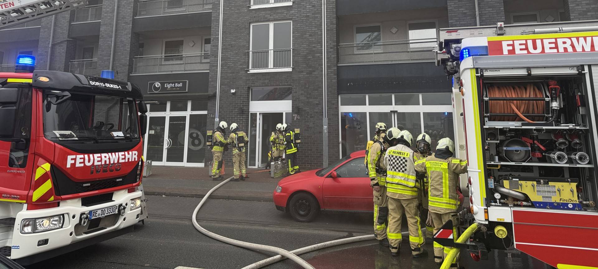 Eine ausgefahrene Drehleiter ist auf ein Wohnhaus gerichtet, aus dem dunkler Qualm aufsteigt. Mehrere Einsatzkräfte und -fahrzeuge sind zu sehen.
